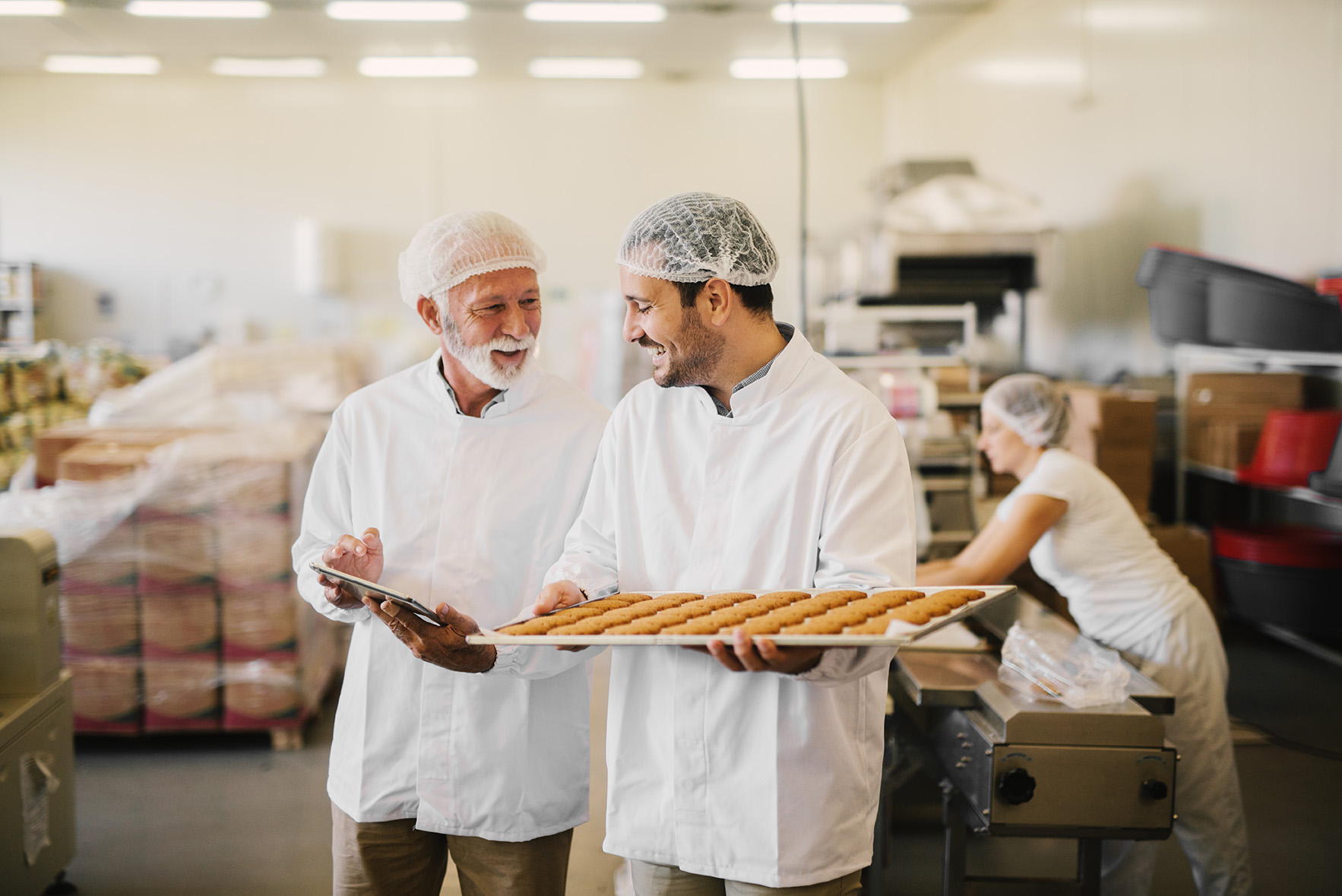 Two men at work talking, one holds a baking try and the other holds a tablet